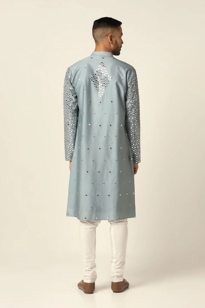 This Chanderi silk Kurta Pajama set features elegant mirror work and thread embroidery on the grey Kurta, paired with off-white churidar pajamas for a timeless look.