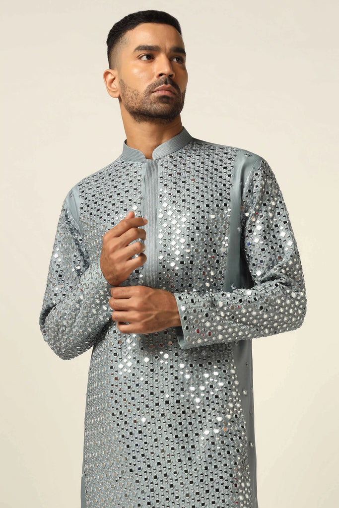 This Chanderi silk Kurta Pajama set features elegant mirror work and thread embroidery on the grey Kurta, paired with off-white churidar pajamas for a timeless look.
