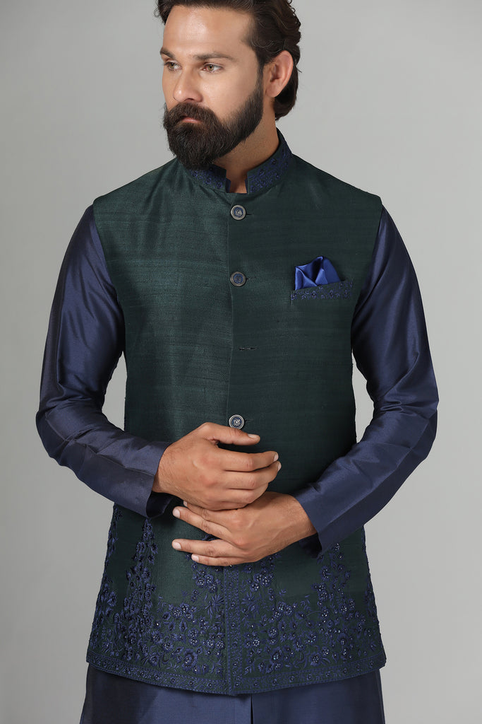 Crafted from luxurious raw silk, this green Nehru Jacket exudes sophistication with subtle embroidery detailing on the collar, hem, and pocket bone. Paired with a short kurta and churidar trousers for a refined ensemble.