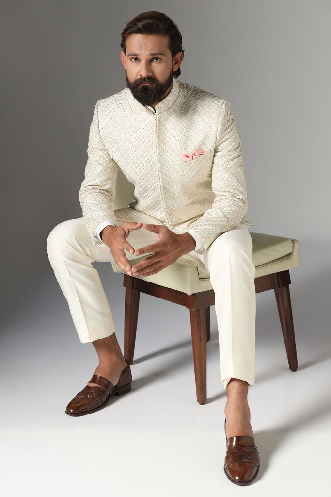 Elevate your style with our off-white Bandhgala suit in raw-silk fabric, featuring zig-zag embroidery. Trousers boast a tapered, flat-front design for a refined look.